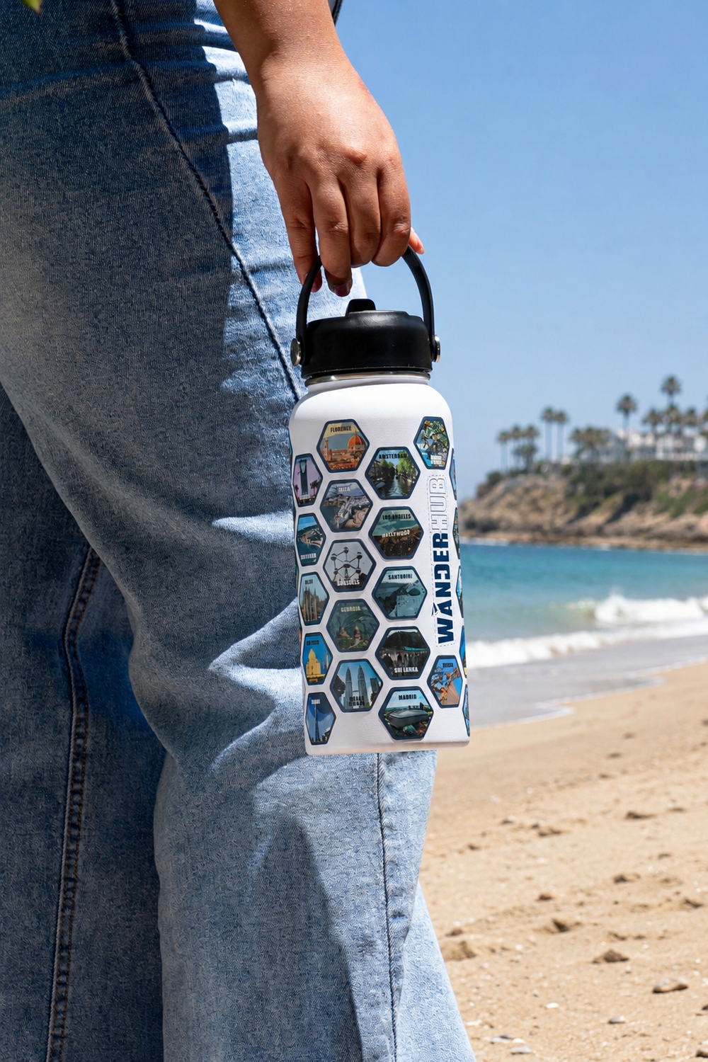 Person holding a travel-themed water bottle with hexagonal pattern on a beach